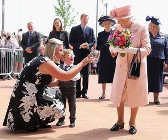 A young boy, visibly upset, meets an elderly woman in a pink outfit holding flowers. A woman is comforting the boy.