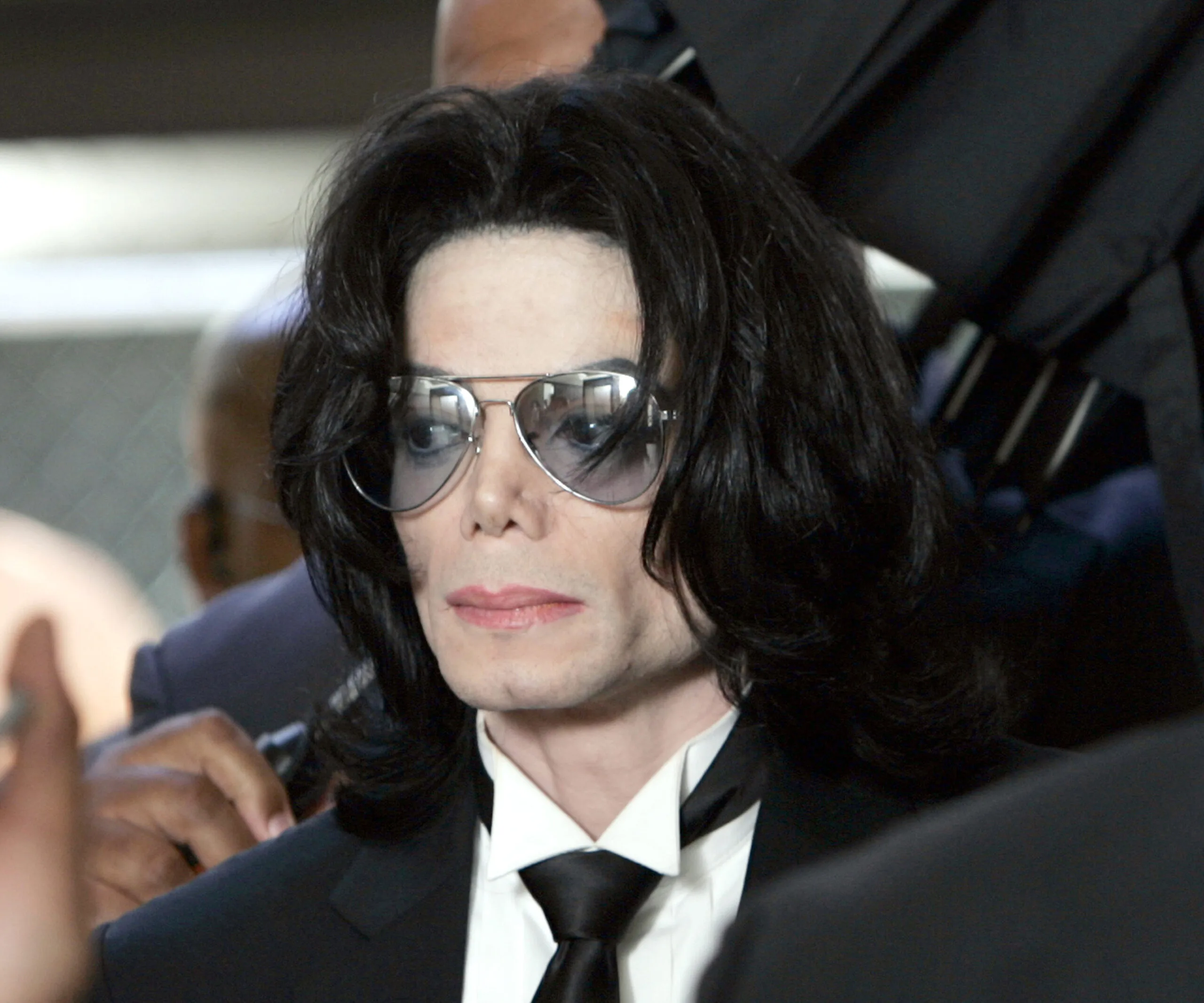 A person wearing sunglasses and a black suit, with long black hair, in a formal setting.