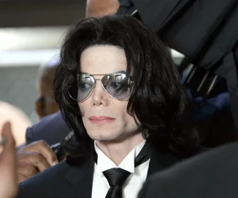 A person wearing sunglasses and a black suit, with long black hair, in a formal setting.