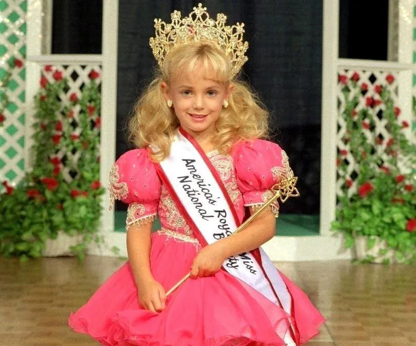 A young girl in a pink pageant dress and crown, holding a scepter, stands on a stage with a sash reading "America's Royal Miss."