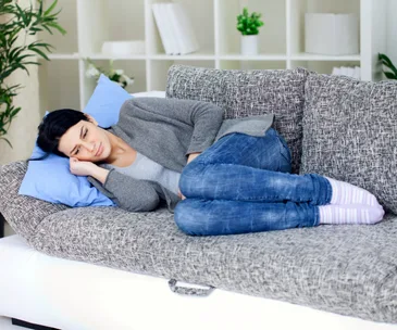 Woman lying on a couch, wearing jeans and a sweater, looking thoughtful and relaxed.