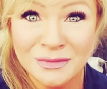 Close-up of a woman with blonde hair and bright eyes, wearing makeup and smiling slightly.