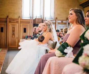 Bride in wedding dress breastfeeding baby during ceremony, seated among guests in a church setting.