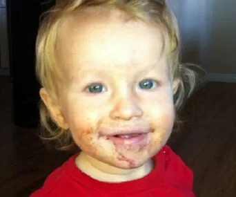 A toddler with blonde hair and blue eyes, wearing a red shirt, and having food stains around the mouth.