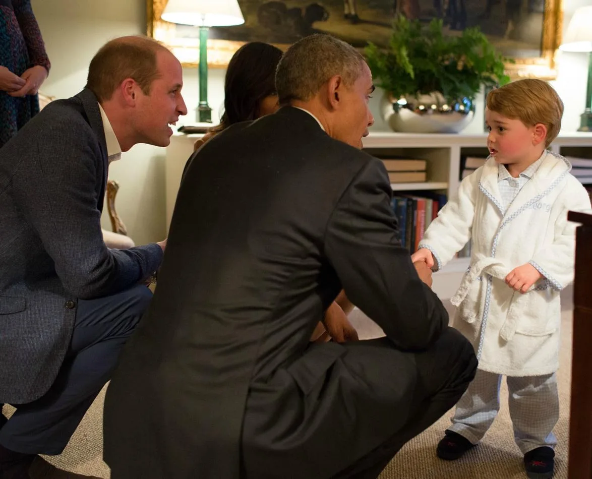 Young boy in a white dressing gown shakes hands with a man in a suit, observed by another man kneeling nearby.