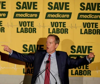 Campaign speaker gesturing in front of "Save Medicare, Vote Labor" signs.