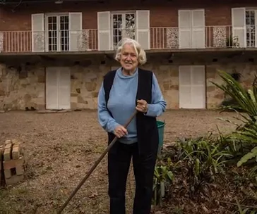 Woman gardening in front of a stone facade house with white shutters.