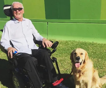 Man sitting in a wheelchair on grass, smiling with a Golden Retriever beside him.