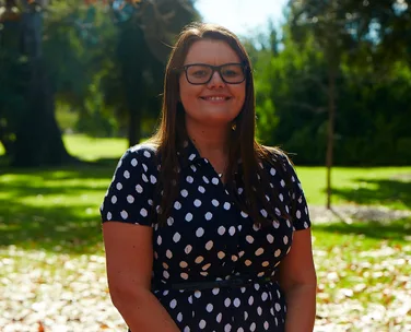 A woman in glasses and a polka dot dress stands smiling in a sunlit park.