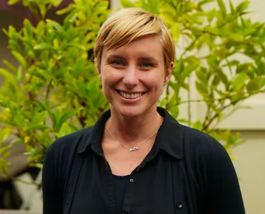 A woman with short blonde hair smiling in front of green foliage, wearing a dark shirt and a necklace.