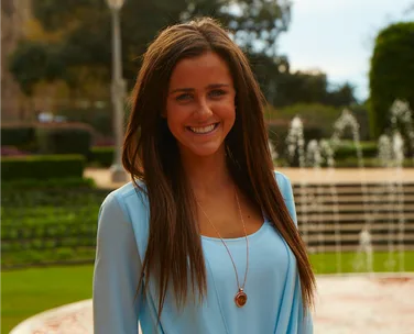Smiling woman in a blue blouse standing outdoors with fountains and greenery in the background.