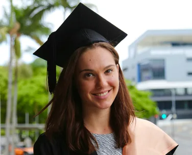A young woman in a graduation cap and gown smiles, with trees and a building in the background.