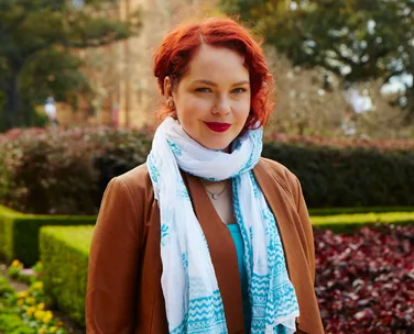 Young woman with red hair and a bright scarf, standing in a garden with hedges and flowers.
