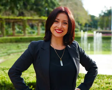 Woman smiling in a garden, wearing a black blazer and necklace, with a fountain and greenery in the background.
