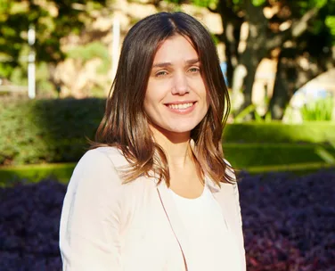 Woman smiling outdoors with trees and shrubs in the background, wearing a light-colored blazer.