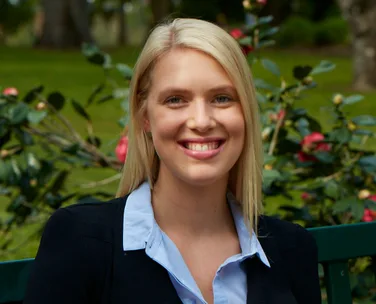 A woman with blonde hair, smiling outdoors in front of flowering bushes.