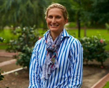 A woman in a blue and white striped shirt with a scarf, smiling in a garden setting.