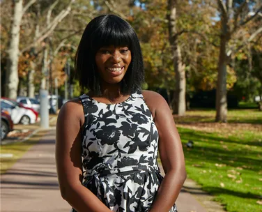 Person smiling in a park, wearing a black and white floral dress, with trees and parked cars in the background.