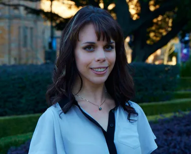 A woman with long brown hair, smiling, in a garden setting.