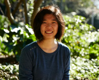 Smiling woman in a blue shirt, seated outdoors with greenery and sunlight in the background.