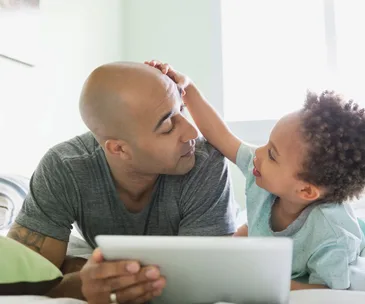 Father and young child smiling and interacting on a bed, holding a tablet.