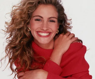 Smiling person in a red sweater with curly hair, embracing themselves against a plain background.