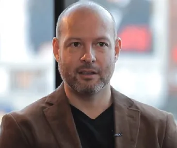 A man with a beard wearing a brown jacket, speaking indoors.