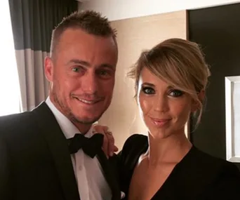 Couple dressed elegantly, posing with smiles in a warmly lit room.