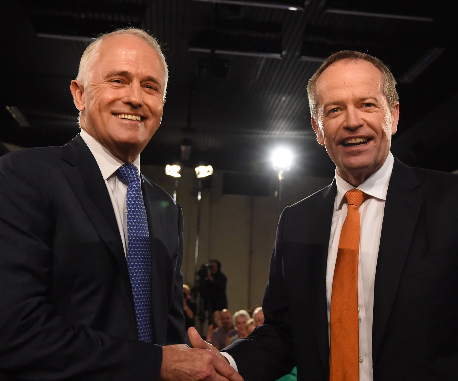 Two men in suits smiling and shaking hands in front of an audience.