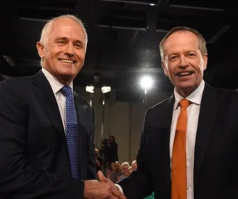 Two men in suits smiling and shaking hands in front of an audience.