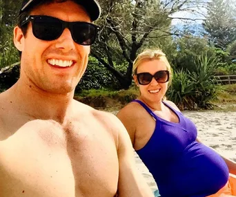 Couple smiling at the beach, woman in a blue top showing her baby bump, both wearing sunglasses.