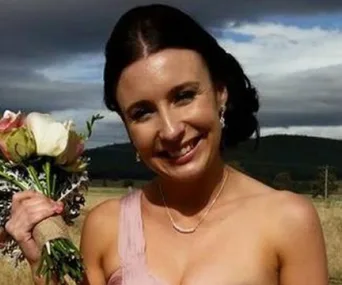 A woman smiling outdoors, holding a bouquet, with hills and a cloudy sky in the background.