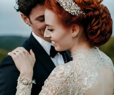 Bride with red hair in ornate gown embraces groom, smiling outdoors.