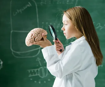A young girl in a lab coat examines a model brain with a magnifying glass in front of a chalkboard.
