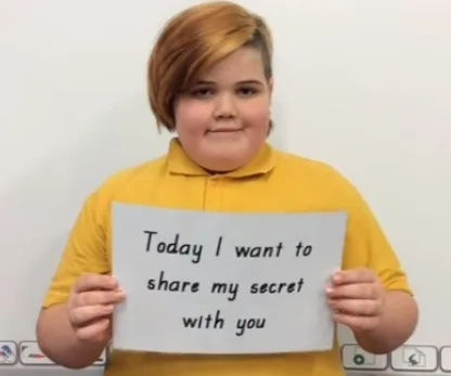 Young individual with short hair holding a sign saying, "Today I want to share my secret with you," wearing a yellow top.