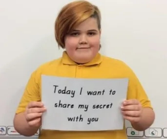 Young individual with short hair holding a sign saying, "Today I want to share my secret with you," wearing a yellow top.