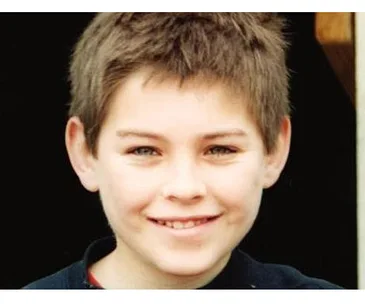Young boy smiling, with short brown hair, wearing a dark top, against a dark background.