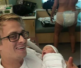 A new father smiles holding a newborn, with a person in a postpartum diaper standing in a hospital room.