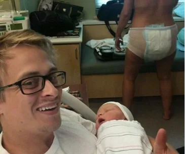 A new father smiles holding a newborn, with a person in a postpartum diaper standing in a hospital room.