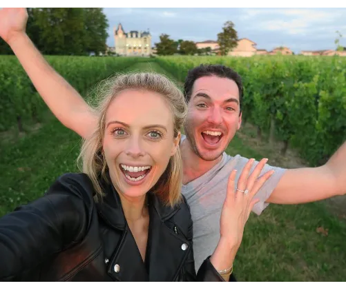 Selfie of a smiling couple in a vineyard with a castle in the background; they appear joyful and excited.