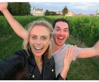 Selfie of a smiling couple in a vineyard with a castle in the background; they appear joyful and excited.