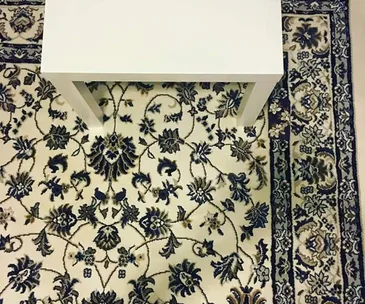 A white table on a patterned carpet with intricate floral designs, designed to camouflage a hidden phone.