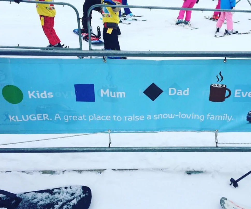 "Banner at a ski resort with symbols for 'Kids', 'Mum', 'Dad', and a coffee cup, promoting Toyota Kluger for snow-loving families."
