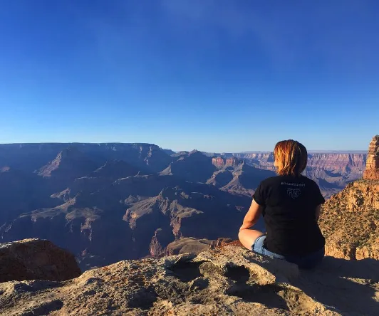 A woman sits on a rock edge overlooking the Grand Canyon under a clear blue sky.