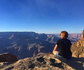 A woman sits on a rock edge overlooking the Grand Canyon under a clear blue sky.