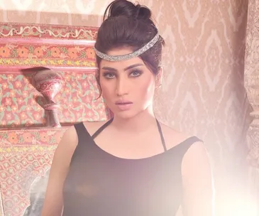 A woman in a black dress with a silver headband stands in front of intricate decor, with soft lighting.