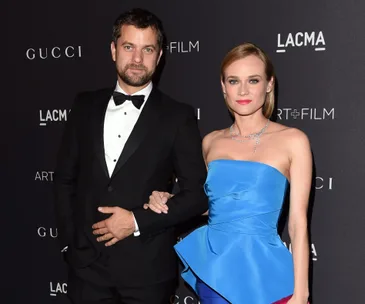 A couple poses together on a black carpet, with him in a tuxedo and her in a blue strapless dress.