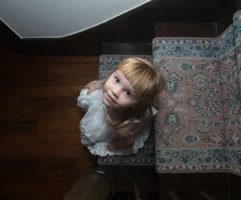 A little girl in a white dress stands on a carpeted staircase, looking up with a curious expression.