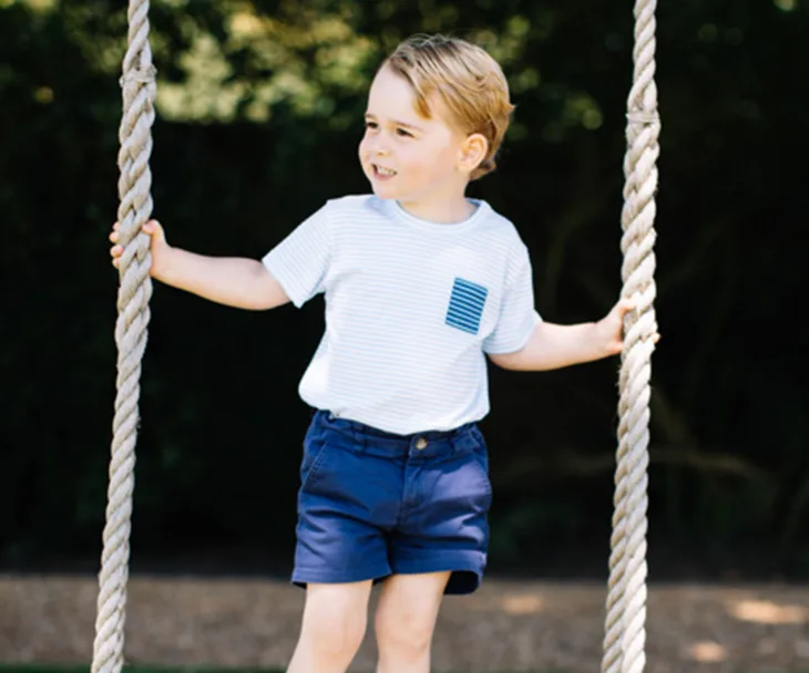 Young child in a striped shirt and blue shorts stands on a rope swing outdoors, smiling and looking to the side.