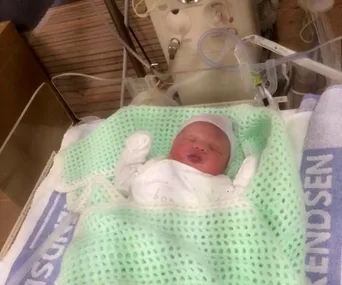 Newborn baby wrapped in a green blanket, lying in a hospital incubator.